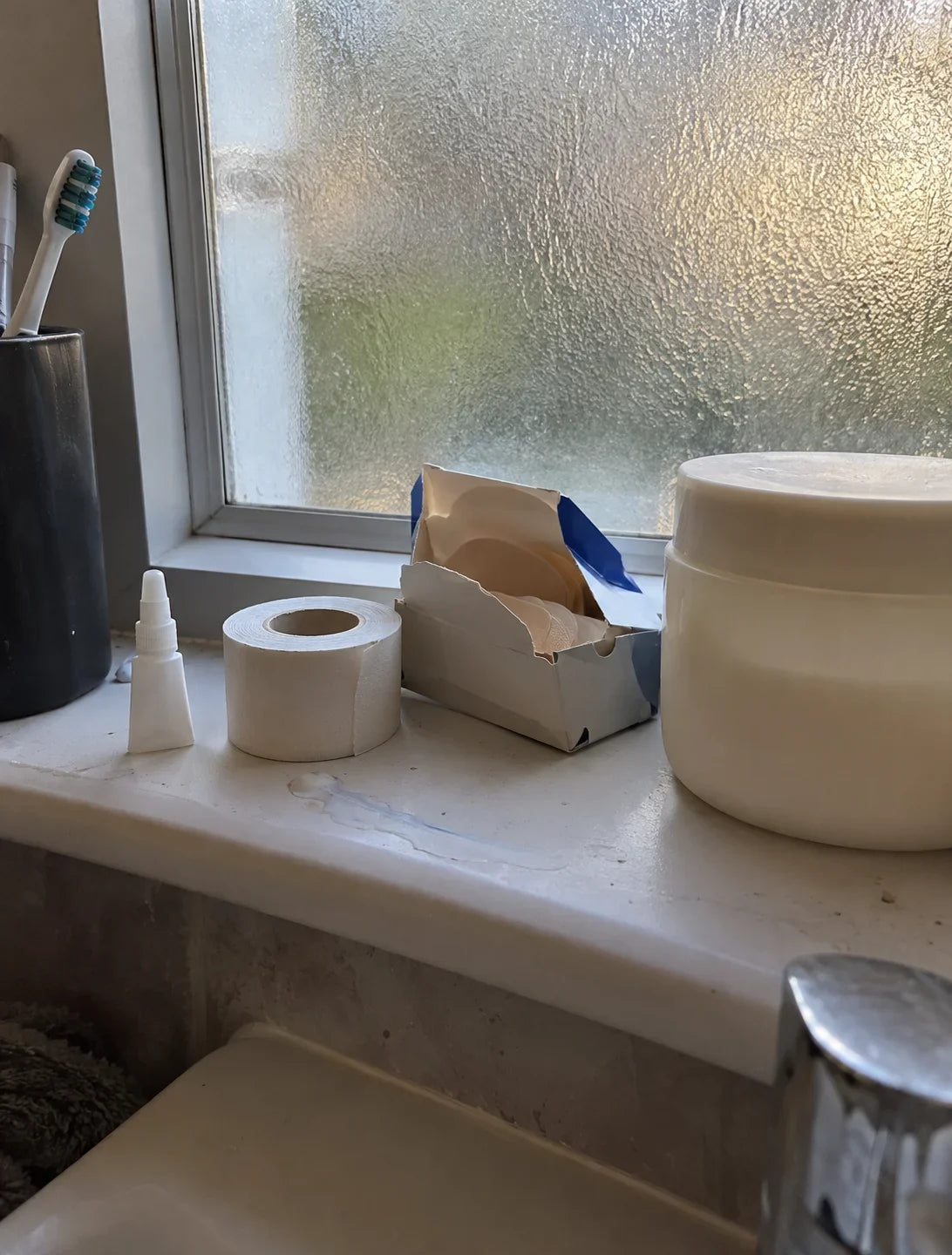 A bathroom windowsill with a tube of superglue, zinc oxide tape, blister plasters and a jar of petroleum jelly