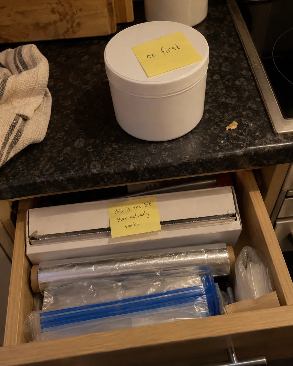 A tub of emollient with a sticky note reading "on first", above an open drawer holding the cling film