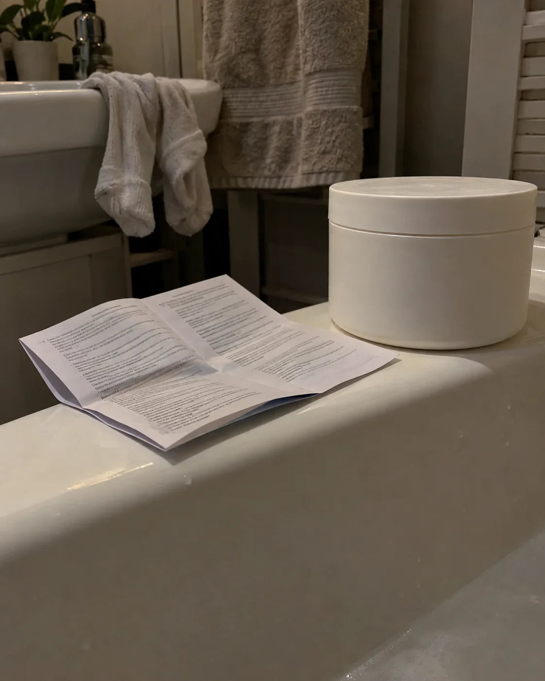 A folded clinic leaflet and a tub of emollient on the edge of the bath, damp socks over the basin behind
