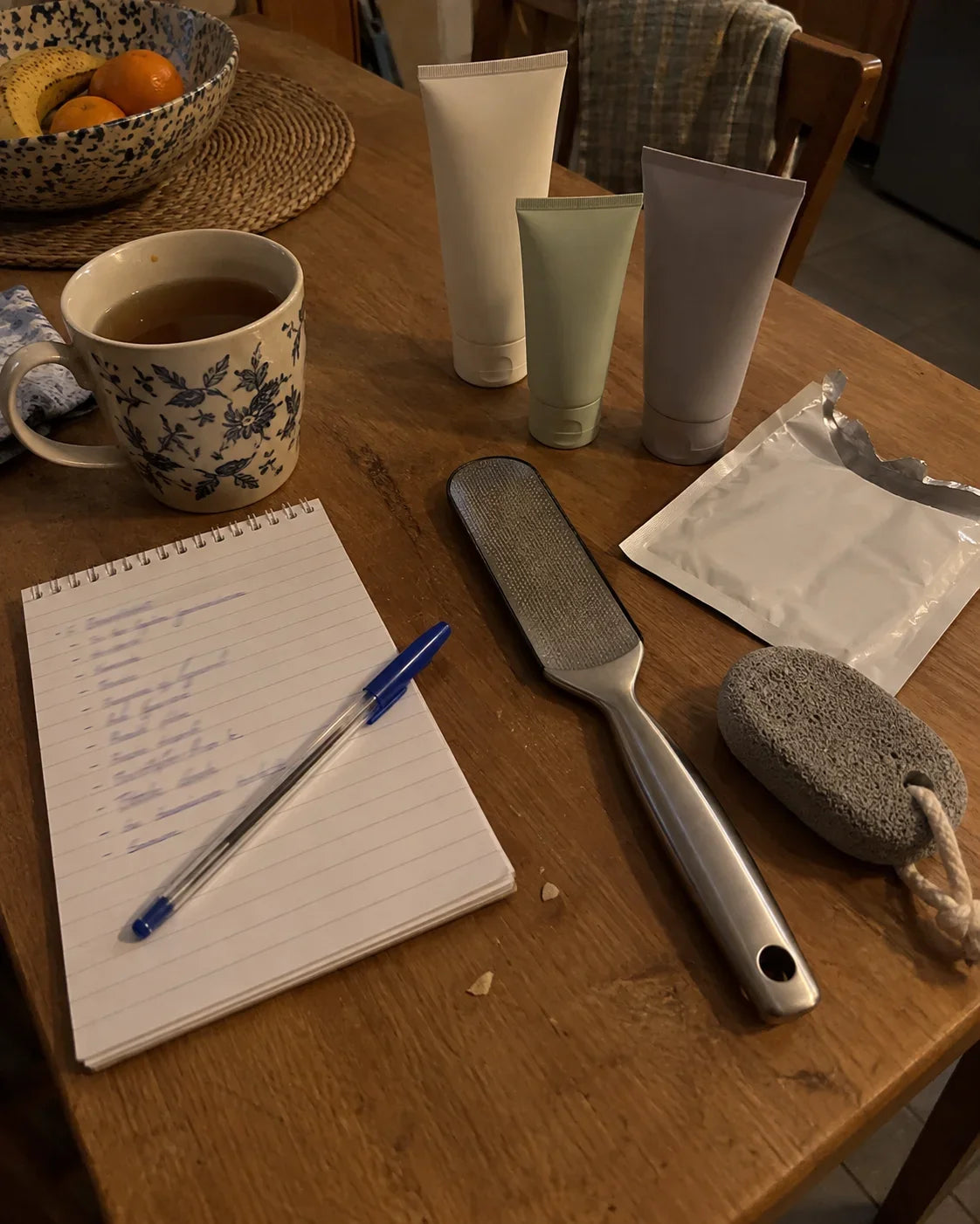 A kitchen table with a mug of tea, a handwritten list, three tubes, a foot file and a pumice stone