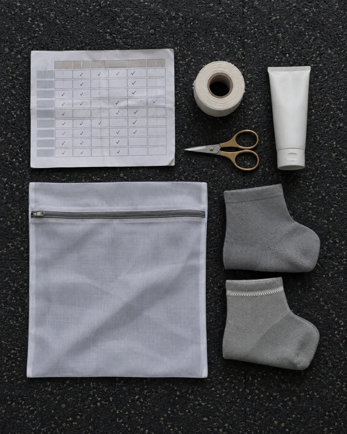 Overhead on a dark rubber floor: a printed training block with ticks, a roll of zinc oxide tape, scissors, a plain white tube, a mesh wash bag and two folded socks