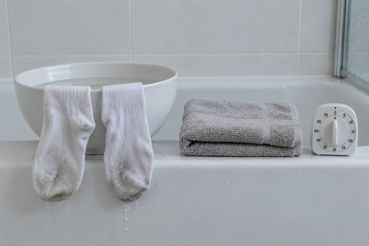 The edge of a bath: a white bowl of water, two damp cotton socks dripping over the rim, a folded towel and a kitchen timer